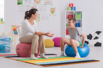 Young physiotherapist and smiling boy sitting on gym ball