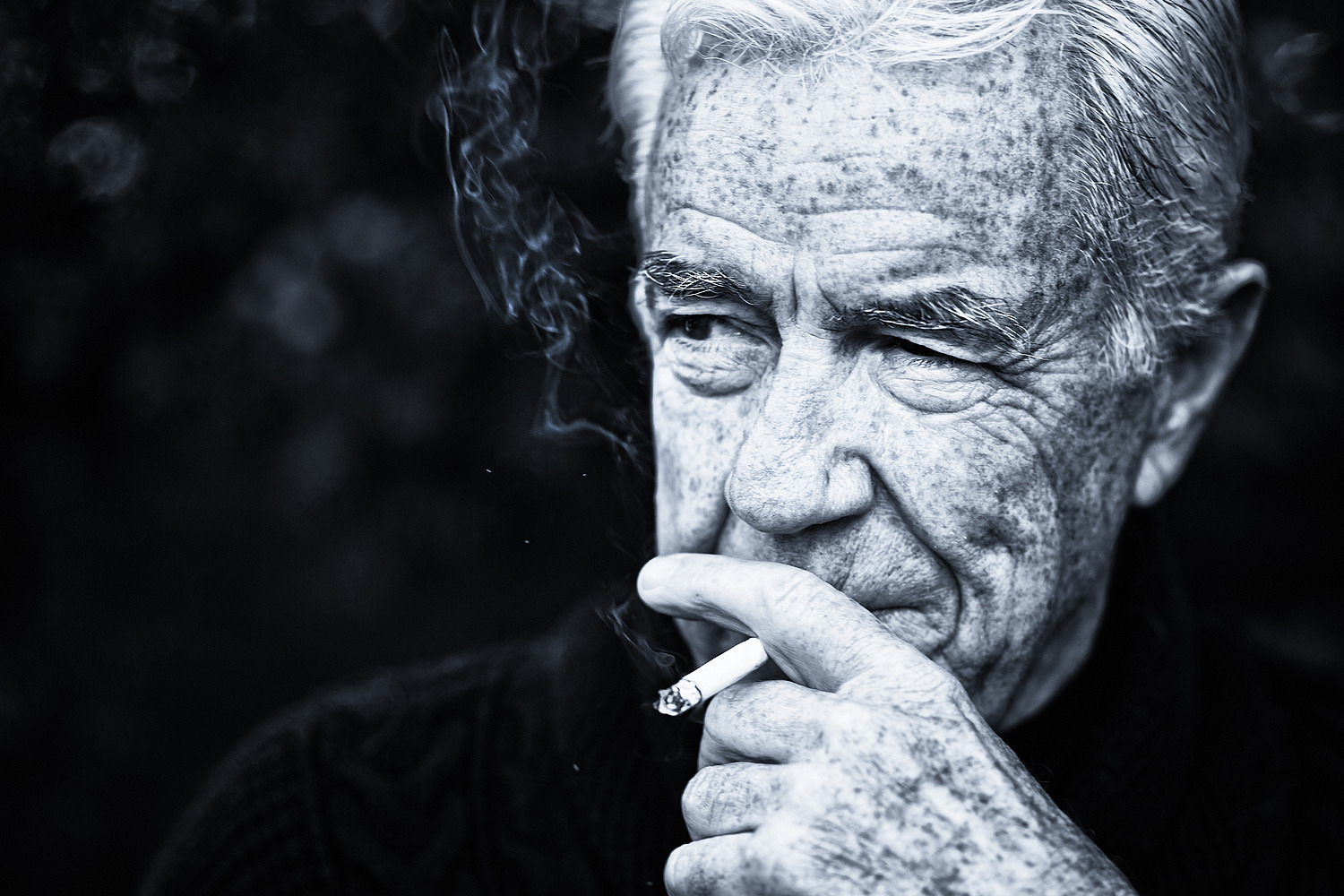 A wrinkled senior man looks sideways as he smokes a cigarette in this monochrome image.