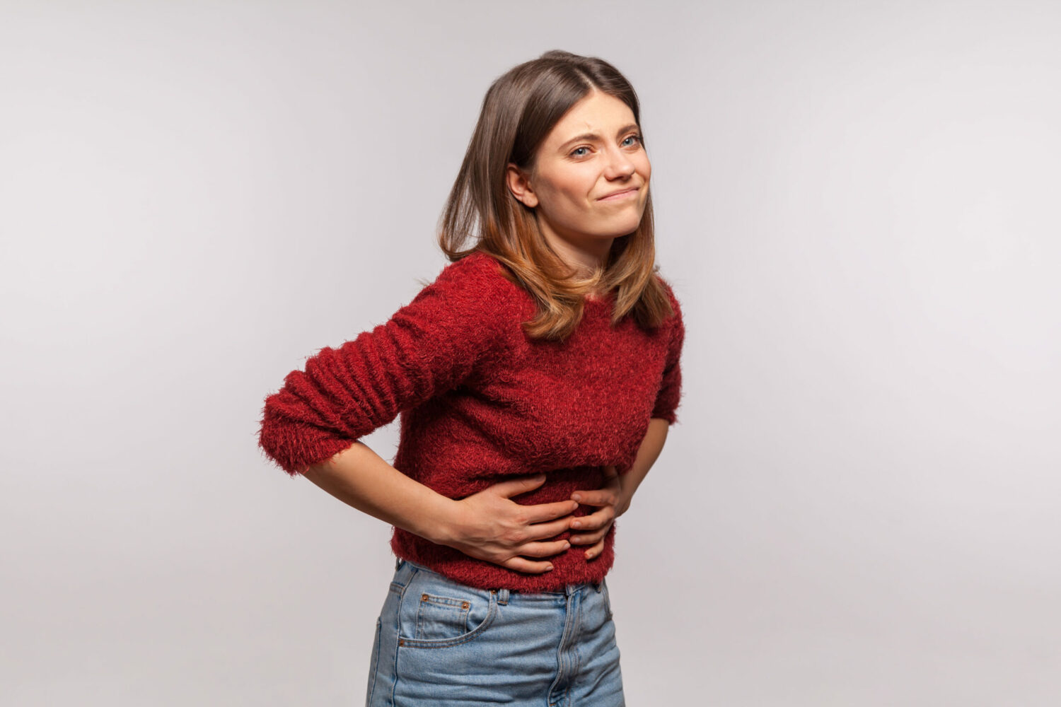 Girl in shaggy sweater touching belly, grimacing from stomach ache, severe abdominal distress, symptoms of constipation, indigestion, gastrointestinal disorder. studio shot isolated on gray background